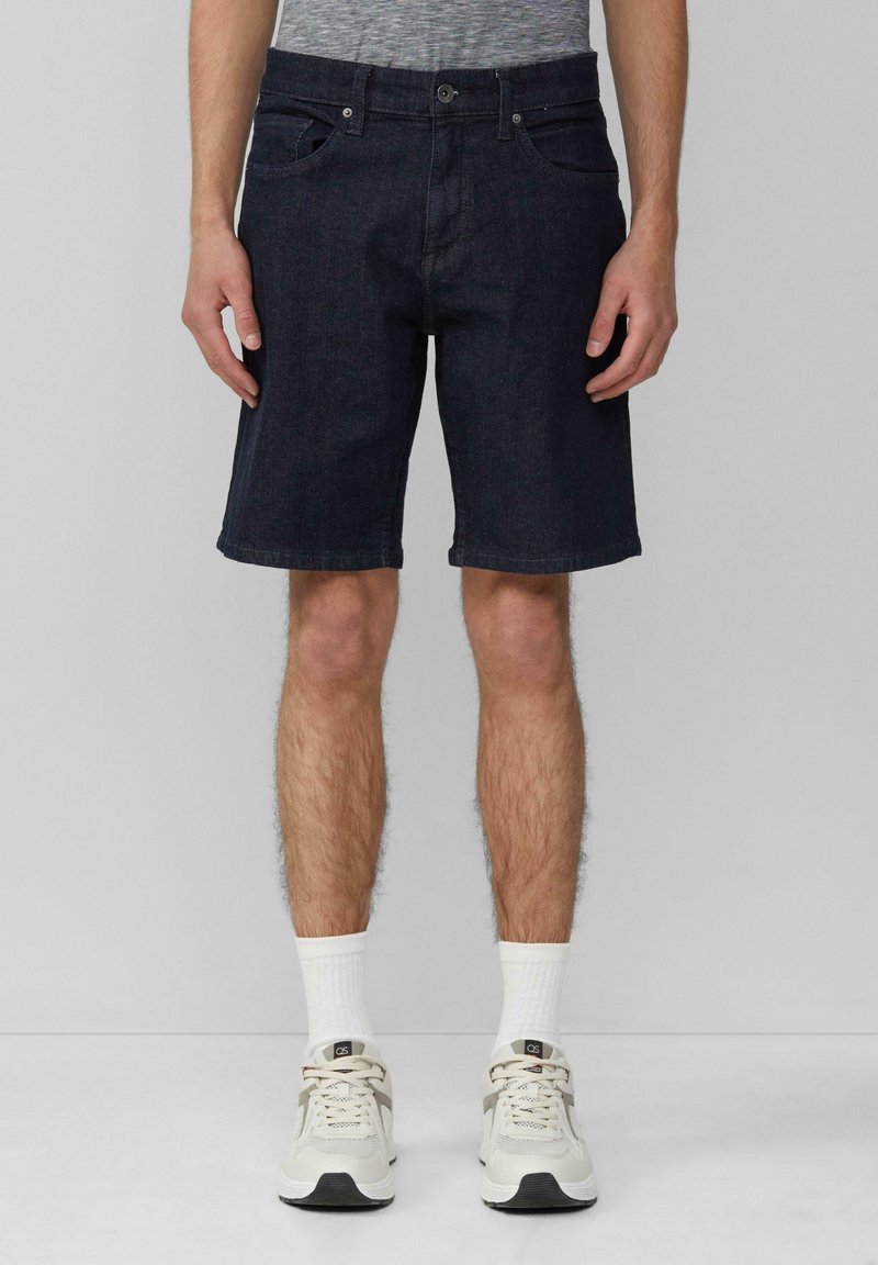 Shorts en denim foncé à coupe classique, avec des poches avant et une fermeture éclair. Portés avec des baskets claires et des chaussettes blanches à la cheville.