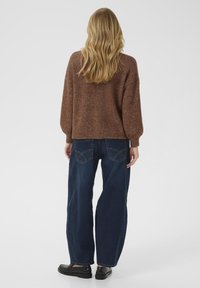 Brązowy, teksturowany sweter dzianinowy z opadającymi ramionami, w połączeniu z luźnymi ciemnoniebieskimi dżinsami z tylnymi kieszeniami i widocznym szwem.