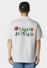 Camicetta bianca a maniche corte con grafiche floreali multicolori e la scritta "PAS DE MONACO" in nero sul retro. Vestibilità casual.