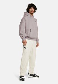 Felpa grigia con cappuccio, grande tasca frontale, abbinata a pantaloni bianchi con dettaglio in zip nera e sneakers nere con accenti bianchi.