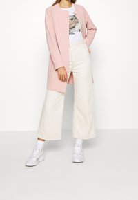 Blazer largo rosa claro, camiseta gráfica blanca y pantalones anchos blancos. Un par de zapatillas blancas completa el atuendo.