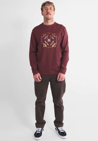 Chemise bordeaux à manches longues avec un imprimé graphique en or et blanc, associée à un pantalon marron foncé et des baskets noires avec des accents blancs.