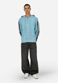 Šviesiai mėlynas megztas užtrauktukas su gobtuvu, juodos ir baltos šoninės juostos, derinamas su plačiomis tamsiai pilkomis džinsais ir baltais sportbačiais.