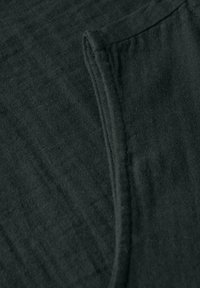 Ciemnozielona tkanina o miękkiej teksturze, z podwiniętym brzegiem i kieszenią. Powierzchnia ma delikatny wzór i gładkie wykończenie.