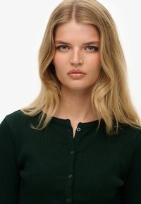 Ciemnozielona prążkowana bluzka z długim rękawem, okrągłym dekoltem i zapięciem na guziki. Miękka faktura, dopasowany krój i subtelny wzór.