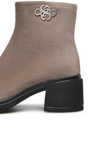 Stivale alla caviglia in suede color taupe con tacco a blocco nero. Presenta un dettaglio decorativo in metallo argentato alla caviglia. Texture liscia.