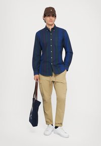 Camisa de botones en azul marino con rayas verdes y moradas; pantalones cargo color caqui; zapatillas blancas; gorra marrón; sosteniendo una bolsa tote azul marino.