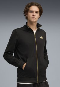Giacca nera con zip realizzata in tessuto liscio, dotata di colletto alto, tasche laterali e logo Puma dorato sul petto.