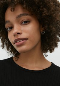 Nahaufnahme einer Person mit lockigem Haar, die einen schwarzen Rippenpullover trägt. Sie hat weiche Haut, minimales Makeup und silberne Creolen.