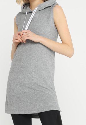 Tunique sans manches gris avec capuche et cordons blancs portée sur des leggings noirs, personne joignant les mains à hauteur de poitrine sur fond blanc.