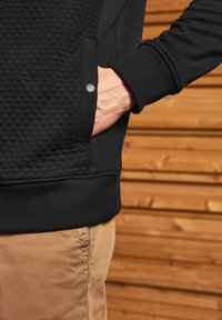Sort sweatshirt med en tekstureret panel og ribbede manchetter, forsynet med en sidelomme lukket med en sort knap, parret med lysfarvede bukser.