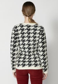 Pulover pufos în carouri alb-negru, cu mâneci lungi și guler rotund. Este purtat cu pantaloni roșii ajustați.