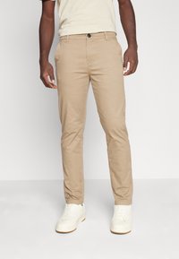 En man som bär beiga raka byxor, off-white sneakers och en kortärmad beige t-shirt står mot en ljus bakgrund.