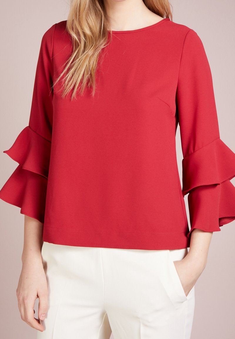 Femme portant un chemisier rouge à manches longues avec des volants étagés sur les manches et un pantalon blanc, debout avec une main dans la poche sur un fond uni.