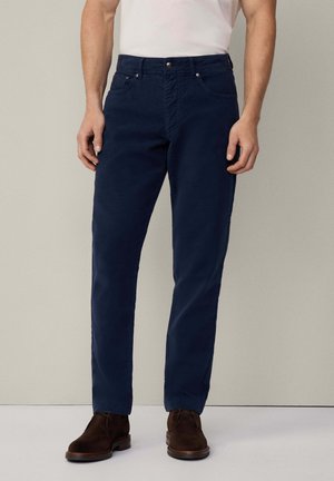 Pantalon en coton bleu marine à coupe droite, avec fermeture éclair, poches avant et texture lisse. Associé à des chaussures en daim marron.