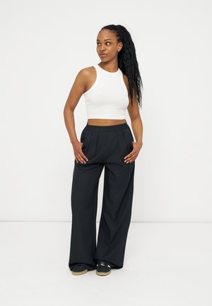 Femme aux longs cheveux noirs tressés, portant un crop top blanc, un pantalon large noir et des baskets noires, debout les mains dans les poches, regardant sur le côté.