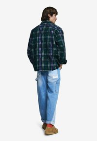 Grünes Karomuster-Fleecehemd mit lila und weißen Akzenten, kombiniert mit hellblauen Cargohosen mit Taschen und aufgekrempelten Bündchen. Braune Schuhe sind sichtbar.