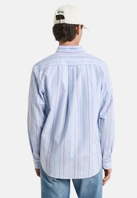Camisa de manga larga de color azul claro con rayas verticales rojas y amarillas, puños abotonados y detalle en el yugo trasero, combinada con vaqueros azules.