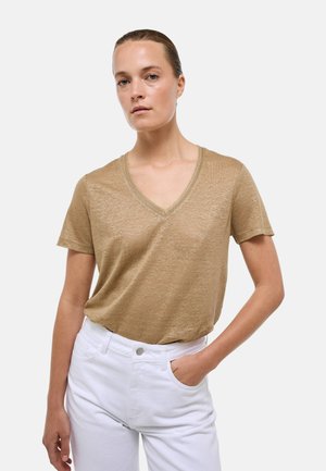 Femme aux cheveux tirés en arrière, portant un t-shirt marron clair à col en V et manches courtes et un pantalon blanc, debout avec une main dans la poche.