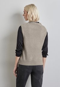 Beige stickad väst med ribbad nederkant, bärs över en svart långärmad skjorta. Mjuk textur, något figurnära silhuett och enkel design.