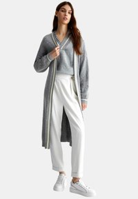 Cardigan grigio lungo con bordi a righe, indossato sopra un maglione grigio abbinato, abbinato a pantaloni bianchi e sneakers bianche.