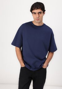 Tricou supradimensionat de culoare albastru marin, fabricat din bumbac, cu guler rotund și mâneci scurte, asociat cu blugi negri. Design simplu, fără modele.
