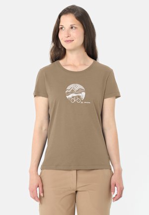 Donna che indossa una maglietta marrone con una grafica circolare di un paesaggio montano e una bicicletta, abbinata a pantaloni marrone chiaro, in piedi davanti a uno sfondo bianco.