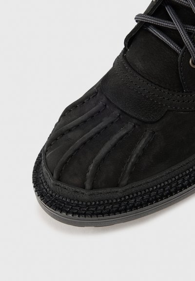 Chaussure noire avec un dessus texturé, présentant des accents cousus et une bordure tissée le long de la semelle. Lacets dans une couleur sombre contrastante.
