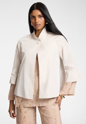 Donna con capelli lunghi neri indossa una giacca crema con bottoni sopra una maglia e pantaloni beige e rosa con motivo, accessoriata con orecchini e bracciale dorati.