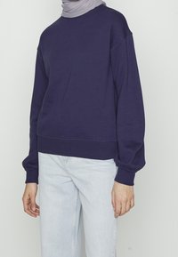 Osoba na sobě má světle šedý hidžáb, tmavě modrou mikinu s dlouhým rukávem a světle modré džíny na jednotném světlém pozadí.