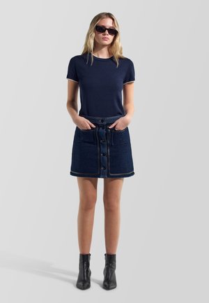 Donna che indossa occhiali da sole neri, camicia blu navy a maniche corte, gonna di denim scuro con bottoni sul davanti e stivaletti neri, in piedi con le mani nelle tasche.