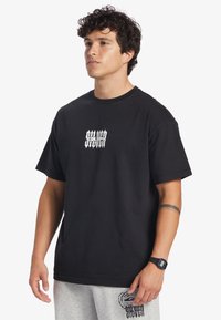 Black cotton t-shirt featuring a small white graphic on the chest, round neckline, and short sleeves. Worn with grey sweatpants.