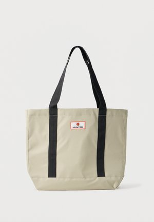 Bežna torba za plažu s crnim trakama. Izrađena od izdržljivog materijala. Ima pravokutan oblik i crvenu i bijelu oznaku s natpisom "HUNTER."