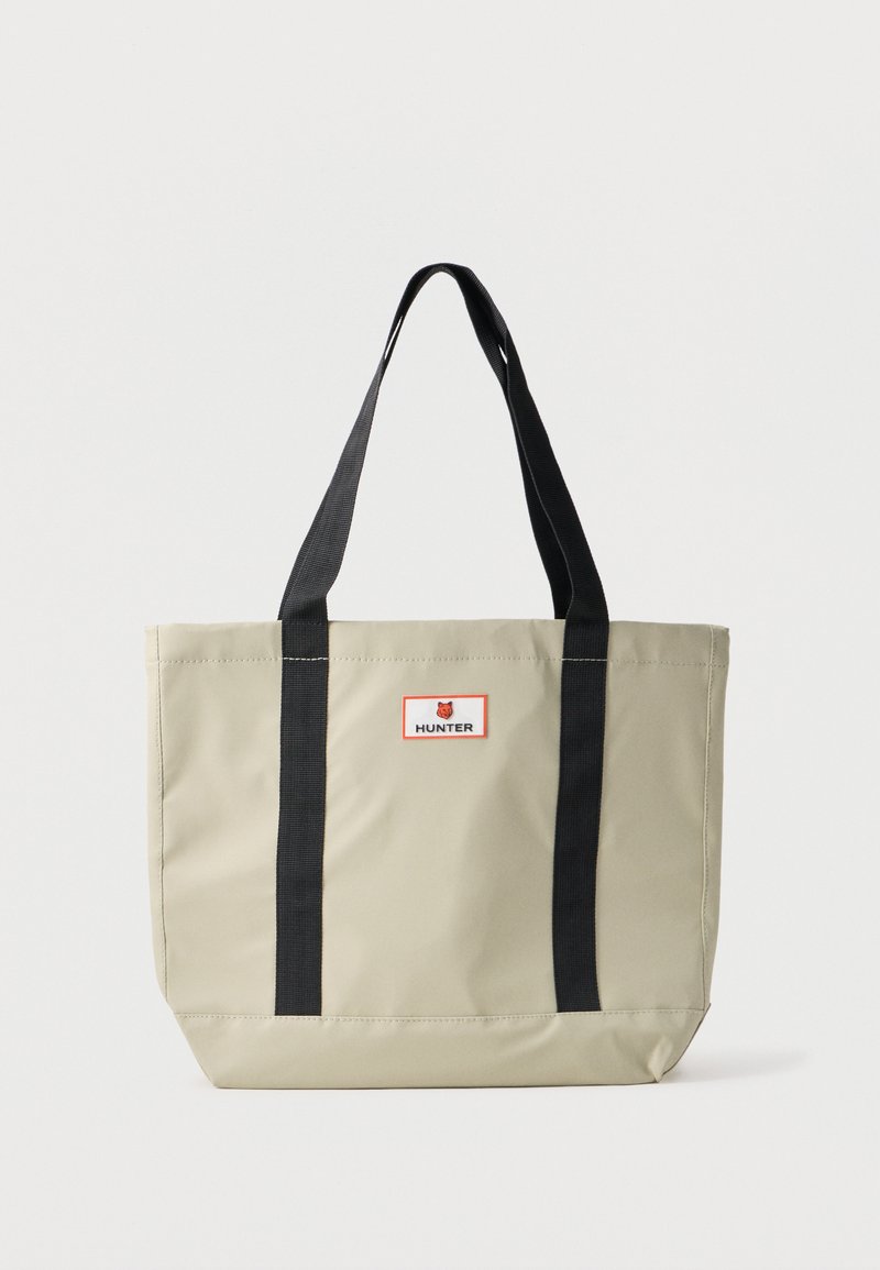 Saco tote bege com alças pretas. Feito de material durável. Apresenta uma forma retangular e uma etiqueta vermelha e branca com "HUNTER."