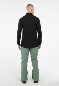 Čierna košeľa s dlhými rukávmi a zelené lyžiarske nohavice s zipsami na členkoch, ktoré má na sebe osoba pohľadom zozadu.