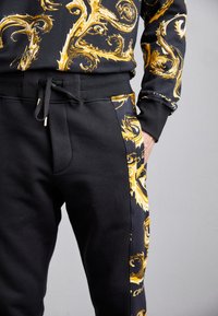 Melna sporta krekls ar zeltainām vijīgām uzdrukām, kopā ar melnām treniņbiksēm, kas rotātas ar zeltainiem akcentiem. Abiem ir ikdienišķs, ērts piegriezums.