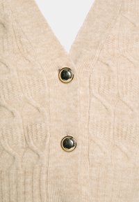Beige knit cardigan featuring a V-neck, textured cable pattern, and two round black buttons with gold accents at the front.