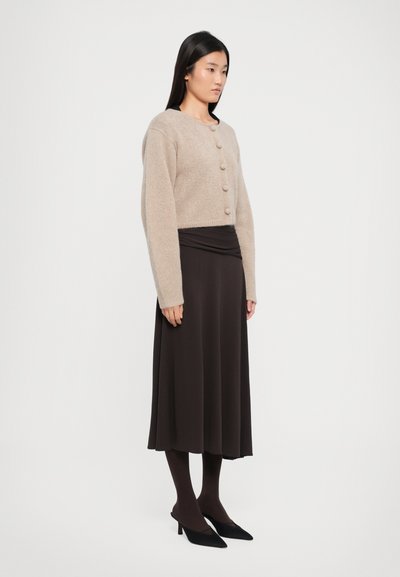 Beige knitted cropped cardigan with round buttons, paired with a dark brown ankle-length skirt and black pointed-toe heels.