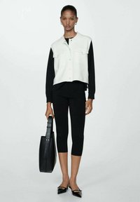 Hvid cropped vest over en sort langærmet skjorte, parret med sorte leggings og spidse sko. Holder en sort tote bag. Enkel design.