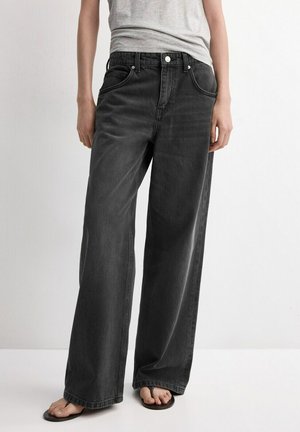 Personne portant un jean noir ample, un t-shirt gris ajusté et des tongs noires, debout devant un fond blanc uni.