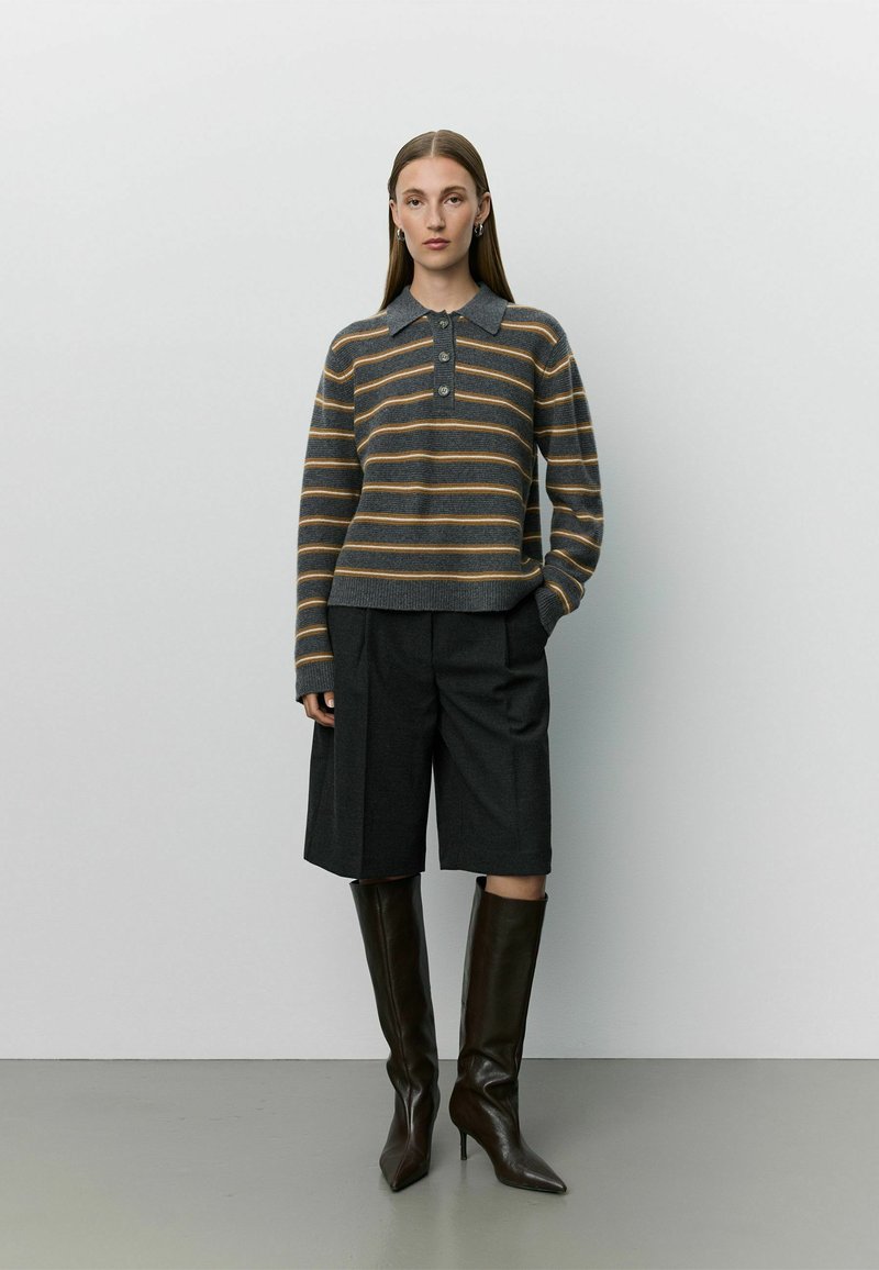 Femme portant un pull gris rayé à manches longues, un short noir arrivant aux genoux et des bottes noires à talons hauts montant jusqu'aux genoux, debout contre un mur uni.