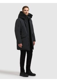 Polstret sort parka med høj krave, lynlås og knaplukninger, flere lommer og et strømlinet design. Båret med sorte bukser og fodtøj.