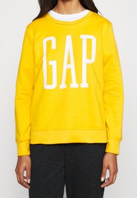 Felpa gialla con logo "GAP" bianco, realizzata in tessuto morbido, con collo a giro e polsini a costine, abbinata a pantaloni neri con motivo.