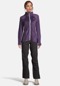 Giacca a zip viola con tessuto testurizzato, colletto alto e design bicolore. Abbinata a pantaloni neri e stivali beige.