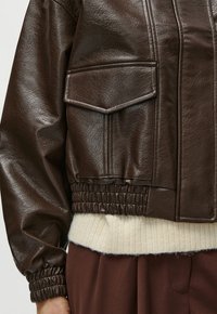 Nahaufnahme einer dunkelbraunen Lederjacke mit großer Klapptasche, elastischen Bündchen und Bund über einem cremefarbenen Strickpullover und braunen Hosen.