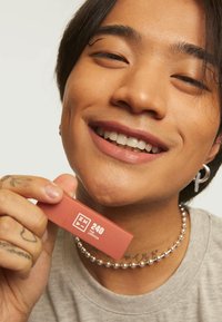 Matt myk ferskenleppeprodukt holdt i hånden, rektangulært emballasje med hvit logo og nummer 240. Modell med kort mørkt hår som smiler.