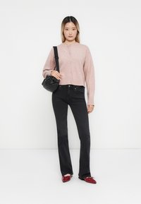 Ljusrosa cardigan med knäppning framtill, i kombination med smala svarta jeans. Hon håller en liten svart handväska och har på sig röda skor.