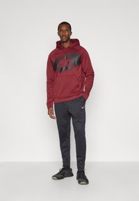 Vyras stovi su rankomis kišenėse, vilkėdamas bordo spalvos "Nike" megztinį, juodas "Nike" treningo kelnes ir juodus sportinius batus prieš paprastą foną.