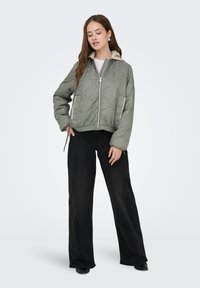 Oliivinvihreä tikattu bomber-takki, jossa on beige kaulus, hopea vetoketju ja sivutaskut, yhdistettynä leveälahkeisiin mustiin housuihin.