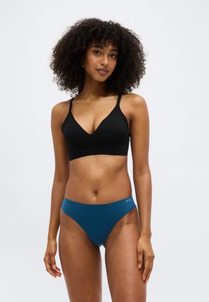 Γυναίκα με σγουρά μαλλιά φοράει μαύρο μπραλέτα και τιρκουάζ εσώρουχο ψηλής κοπής, στέκεται μπροστά σε απλό λευκό φόντο.